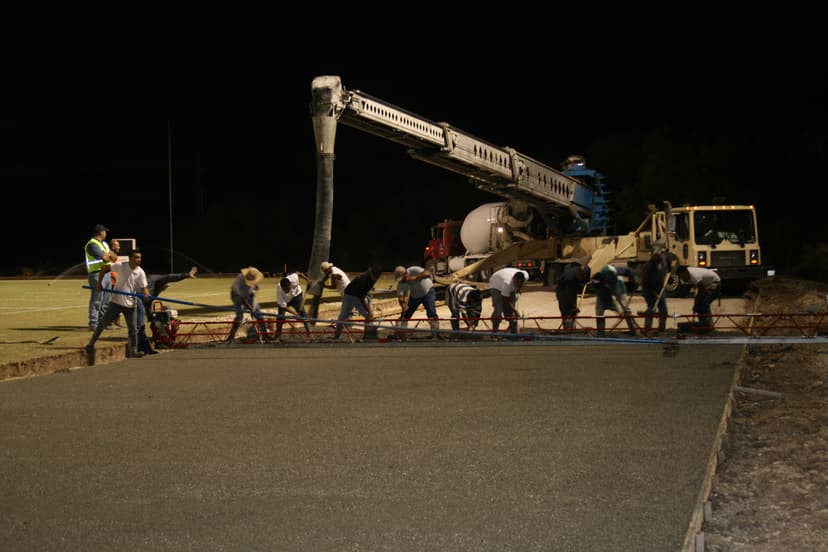 Construction workers pouring concrete at night with a concrete pump and machinery on-site.